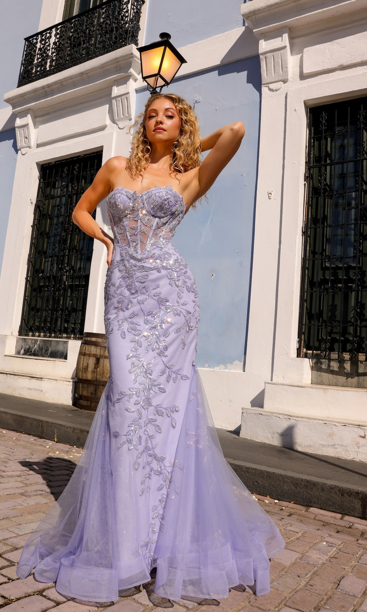 A woman wearing the Sheer-Bodice Strapless Mermaid Prom Dress G1258 with a corset-style bodice and beaded embroidery poses on a cobblestone street in front of a white building with black window bars and a lantern.